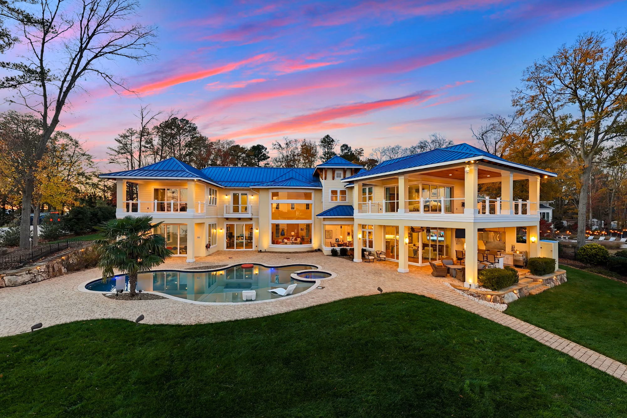 A lakefront house lit from within at dusk, seen from the air above its pool and lawn.