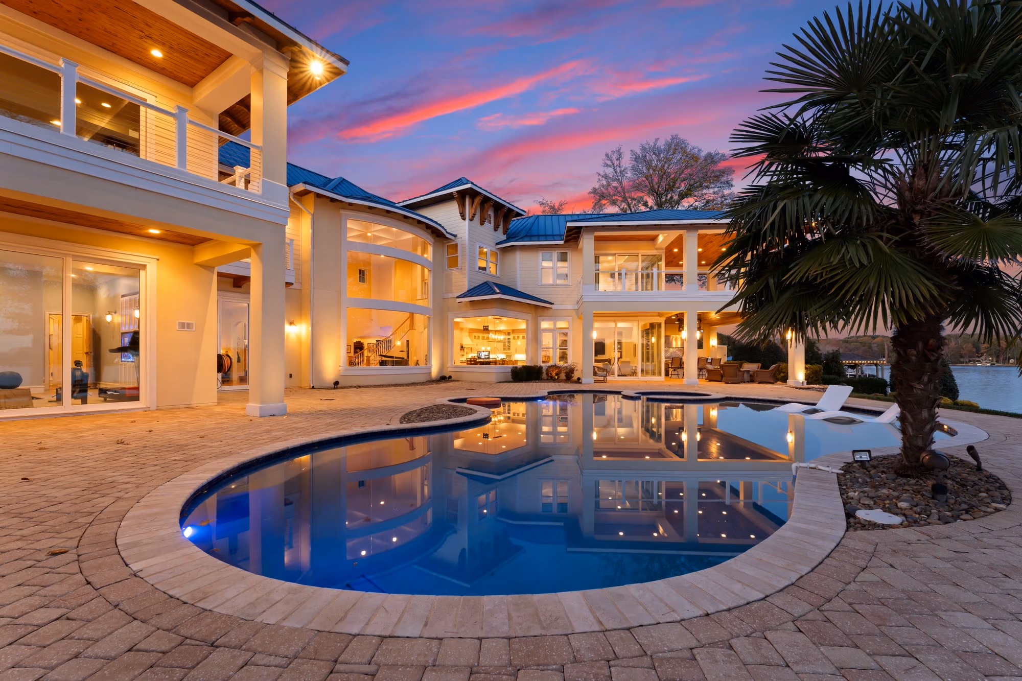 A pool and spa lit blue on a paved terrace at dusk, with the house behind them.