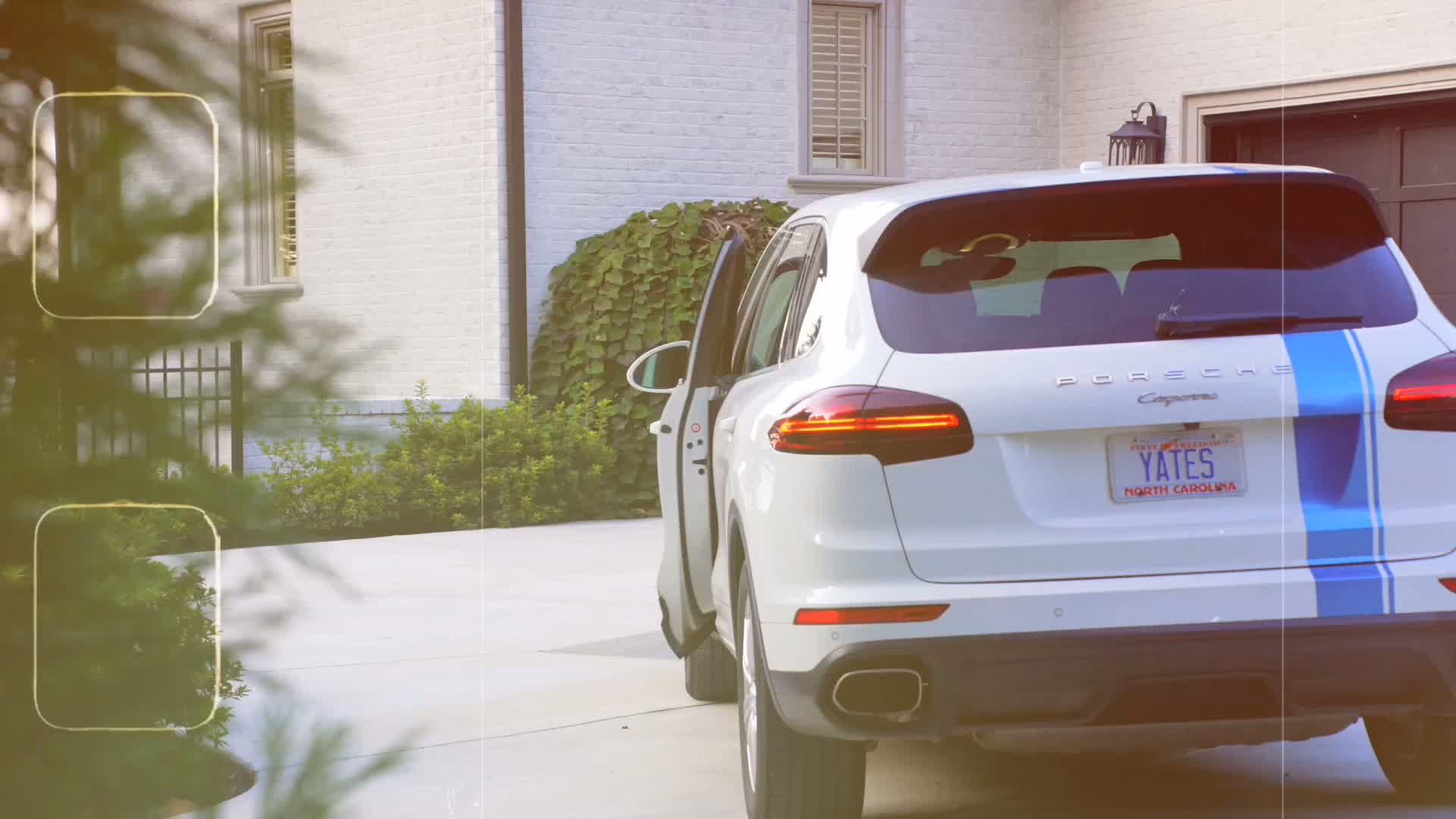 A white SUV with a blue stripe and a YATES North Carolina plate, its door open, in the driveway of a white brick house.