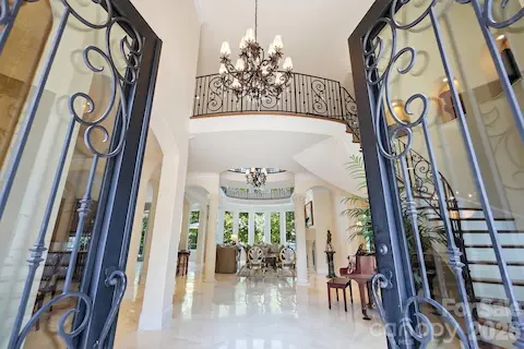 A dramatic first impression begins at the grand two-story foyer, where custom wrought-iron and glass entry doors open to an elegant, light-filled interior. A statement chandelier, sweeping Brazilian cherry staircase, ornate iron railings, soaring ceilings, marble flooring, and graceful architectural columns showcase the exceptional craftsmanship of this Mediterranean-inspired Lake Norman estate. The open sightline through the foyer toward the great room and walls of windows beyond hints at the spectacular waterfront setting that defines the home.
