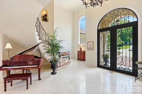 Step through the ornate iron-and-glass double doors into a soaring, light-filled foyer designed to make an immediate impression. Polished stone flooring, a sweeping curved staircase with decorative ironwork, a statement chandelier, and tall arched windows create a refined sense of scale, while the grand piano adds warmth and elegance to the space.