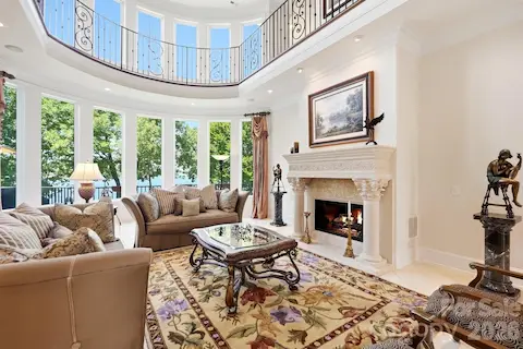 The dramatic two-story great room is framed by a wall of windows overlooking Lake Norman, filling the space with natural light. An ornate fireplace, upper-level gallery, and elegant architectural detailing create a warm yet impressive gathering space.