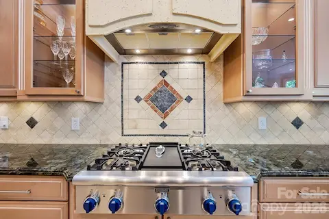 The kitchen’s professional-style range creates a striking focal point, framed by illuminated glass-front cabinetry, a substantial custom hood, and decorative tilework. Rich stone counters and thoughtful detailing give this cooking area a polished, chef-inspired feel.