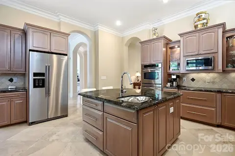 From the opposite side, the expansive center island provides additional prep space, storage, a sink, and convenient seating. The kitchen remains visually connected to the adjoining casual living and outdoor areas, allowing natural light and views of the surrounding greenery to carry through the space.
