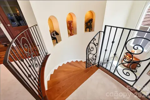 The sweeping staircase continues to the upper level with rich wood treads and custom wrought-iron railings. Illuminated wall niches add architectural interest while the open design maintains a visual connection to the dramatic foyer below.