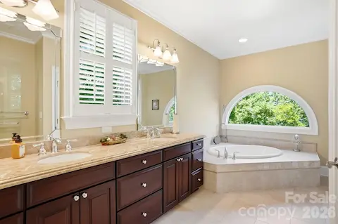 The private bath features a long dual-sink vanity with rich cabinetry and stone countertops, along with a spacious soaking tub positioned beneath a distinctive arched window. Natural light and leafy views give the room a bright, relaxing feel.