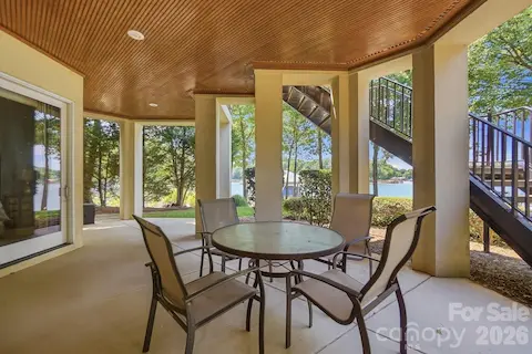 The lower-level living spaces open to a broad covered patio with a warm wood-plank ceiling, recessed lighting, and multiple seating areas. Protected from the elements yet open to the landscaped yard and lake beyond, it extends the home’s entertaining space outdoors.