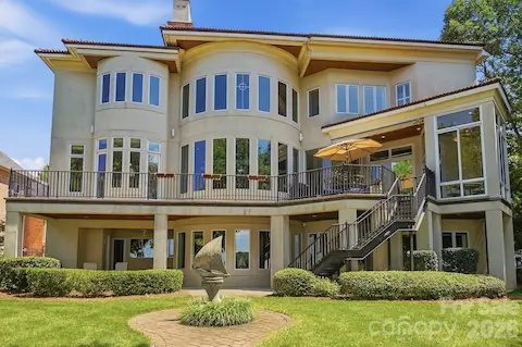 A full rear view showcases the home’s impressive three-level design, expansive windows, upper terrace, covered lower patio, and exterior stair connection between levels. Manicured landscaping frames the residence and highlights the scale of the outdoor living areas leading toward the Lake Norman waterfront.
